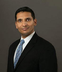 Tomlee L. Abraham. He is a medium-skinned adult man who has black hair. He is wearing a black suit jacket, white collared shirt, and a blue patterned tie.