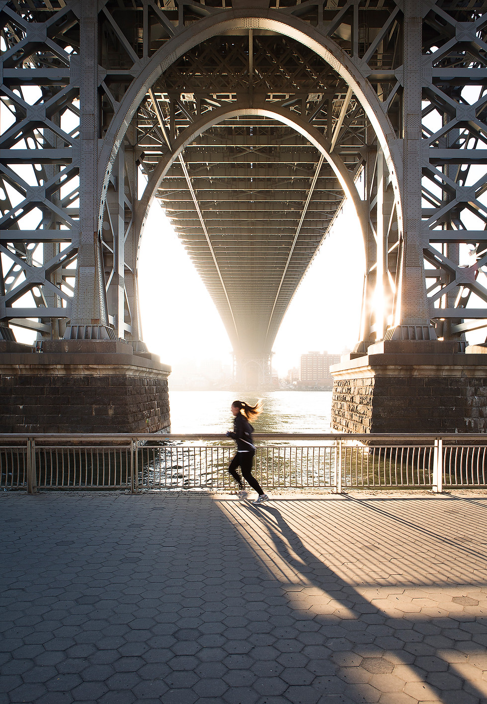 Runner beneath the Williamsburg Bridge in New York, silhouetted against the afternoon sun with long shadows across the waterf