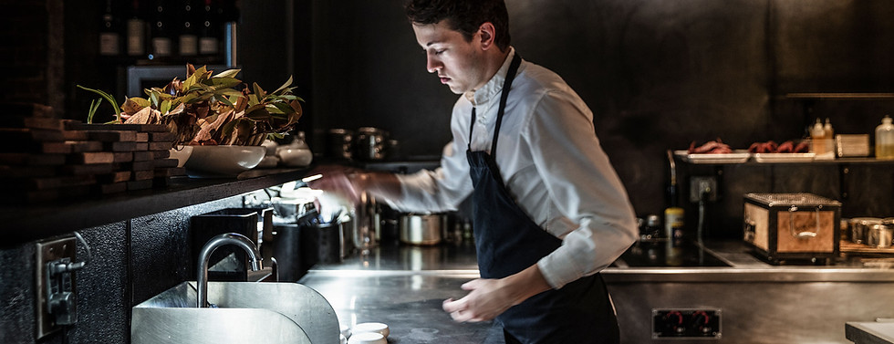 Chef working at a stainless steel counter in the dimly lit kitchen of Restaurant Aska in Brooklyn, New York.