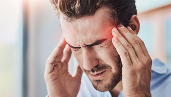 A man presses his temples with both hands, eyes shut in discomfort, showing symptoms of a cervicogenic headache often linked to upper cervical spine misalignment.