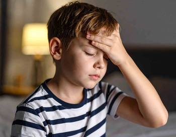 Young boy sitting indoors with his eyes closed and his hand on his forehead, appearing to experience a headache or discomfort.