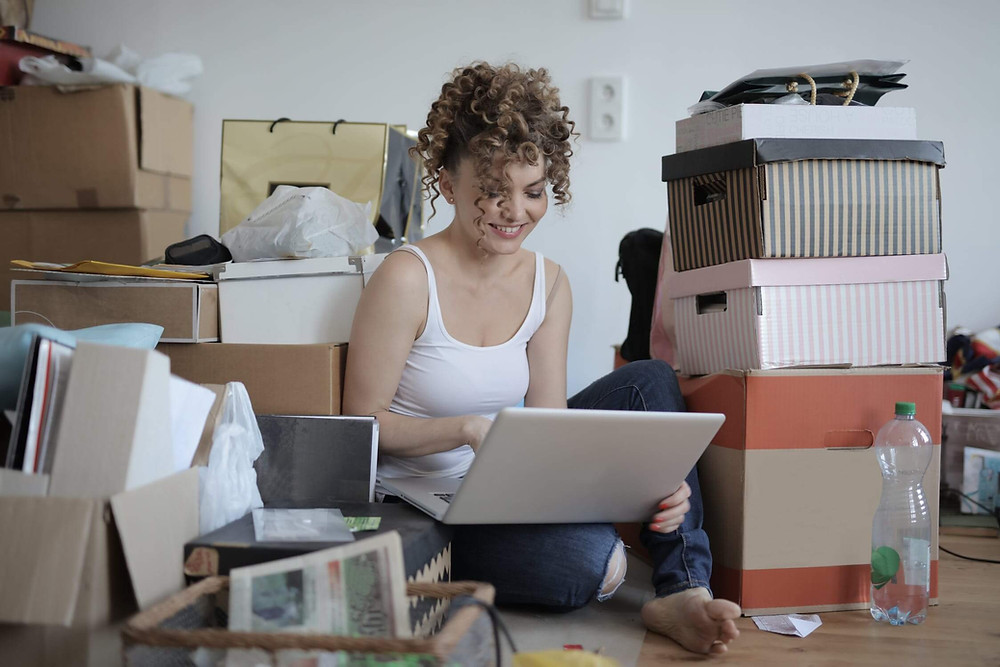 A woman sitting on the floor with her laptop shopping online while surrounded by endless piles of packaging and boxes