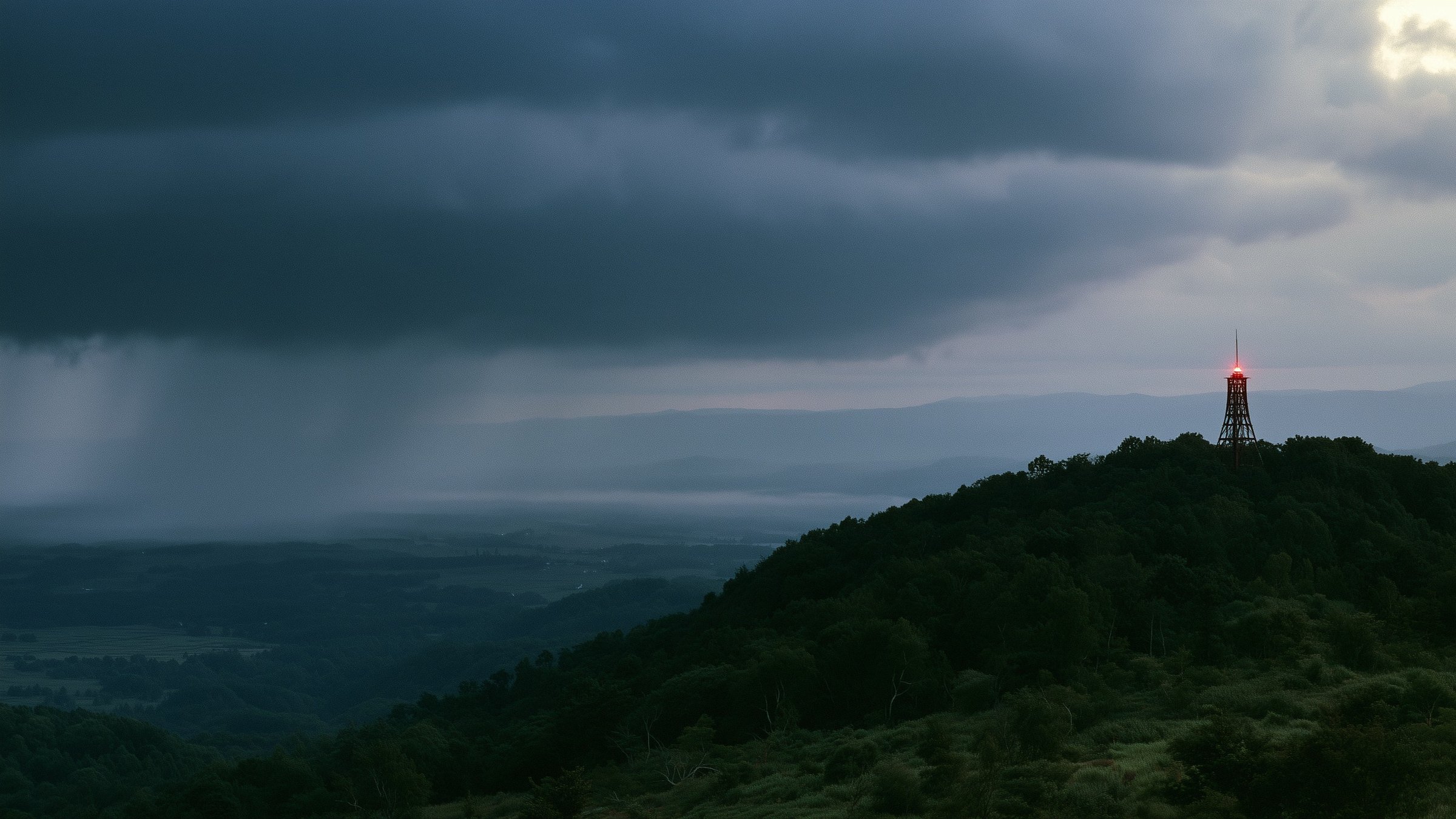 A lattice tower with a red light on a dark hilltop under storm clouds.