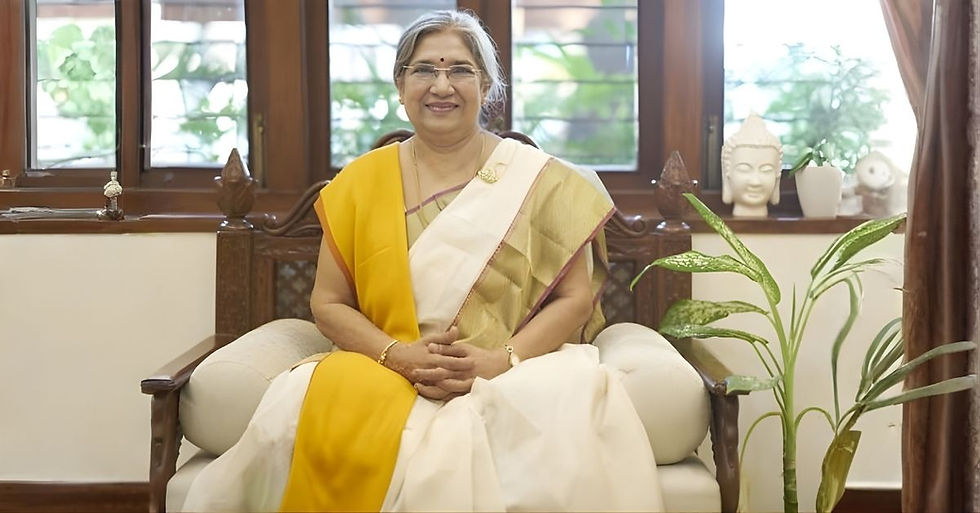 Dr. Hansaji Yogendra, in a yellow and white saree, seated in a wooden chair with a Buddha statue 