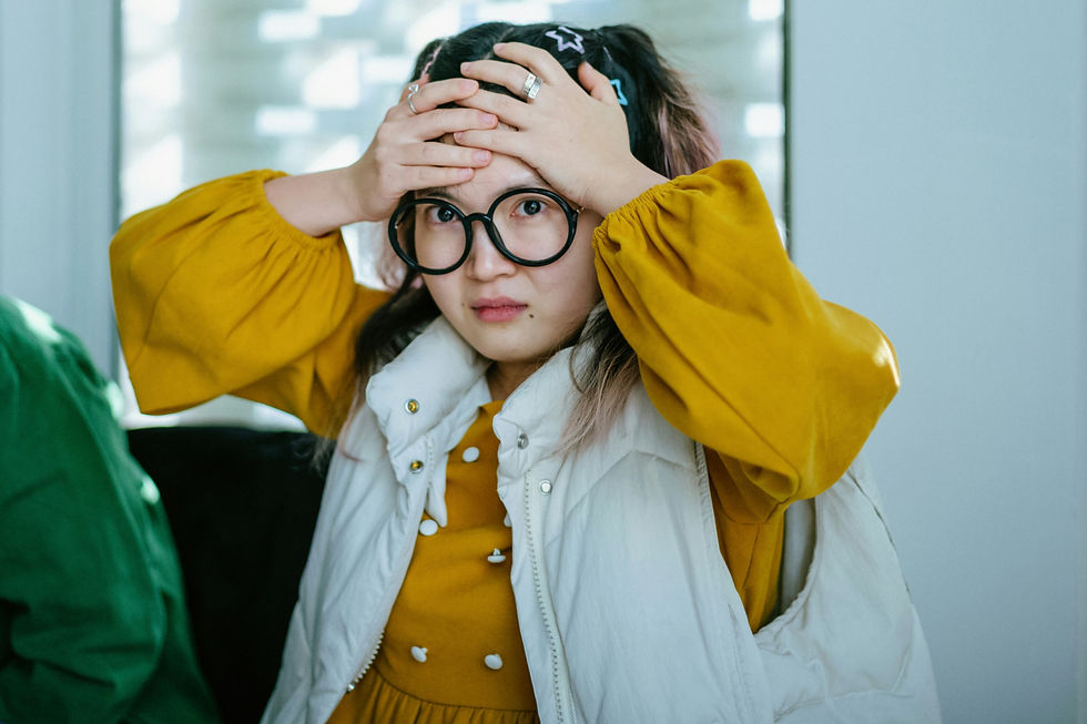A person with large glasses holds their head in concern. They wear a yellow dress and white vest, set against a bright, blurred background.