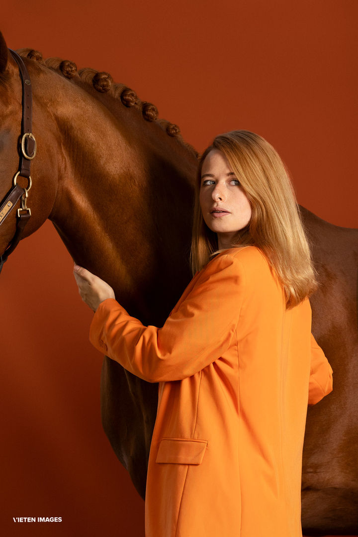 Studio portrait of photographer Diana Wahl and her horse Wolke, wearing an orange suit, posing  in front of an orange background