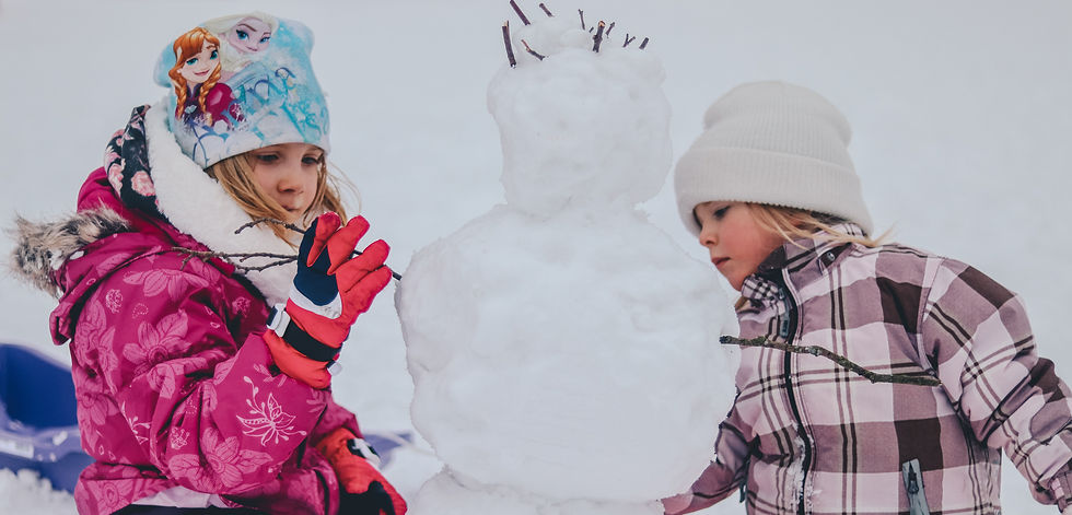 Two children in winter gear building a snowman with sticks. One wears a pink jacket; the other has a plaid coat and white hat. Snowy background.