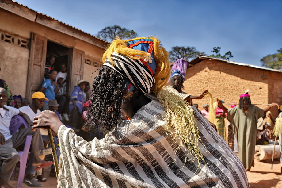 Kpelle person in vibrant traditional attire dances outdoors, watched by a crowd.