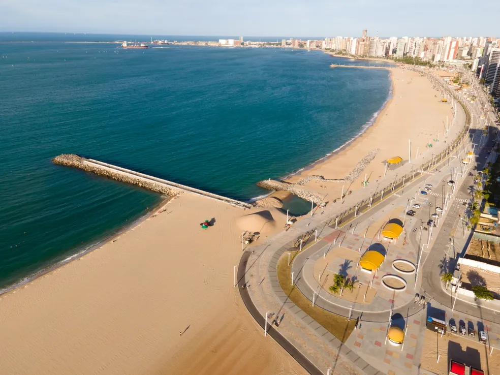 Vue aérienne de la plage d'Iracema à Fortaleza montrant le nouveau remblai, la jetée de pierre et les gratte-ciel du bord de mer sous un ciel bleu.