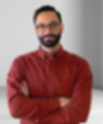 Man with glasses in a red patterned shirt, arms crossed. He stands in a minimalist room with a gray gradient wall, appearing confident.