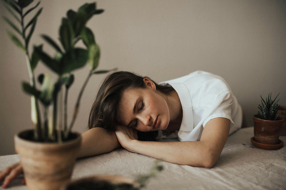 Woman in white shirt resting her head on a table, surrounded by potted plants. The setting is calm and earthy, evoking a serene mood.