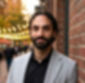 Man with a beard in a gray blazer smiles slightly, leaning against a brick wall. Blurred city lights and autumn trees are in the background.