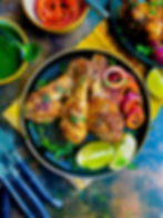 Grilled chicken legs on a black plate with lime and salad. Green sauce and sliced red onions in bowls. Rustic table setting.