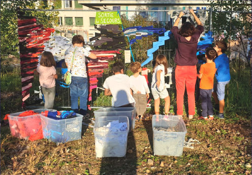 Une dizaine de personnes de dos en extérieur, elles attachent des bouts de tissus sur une barrière.