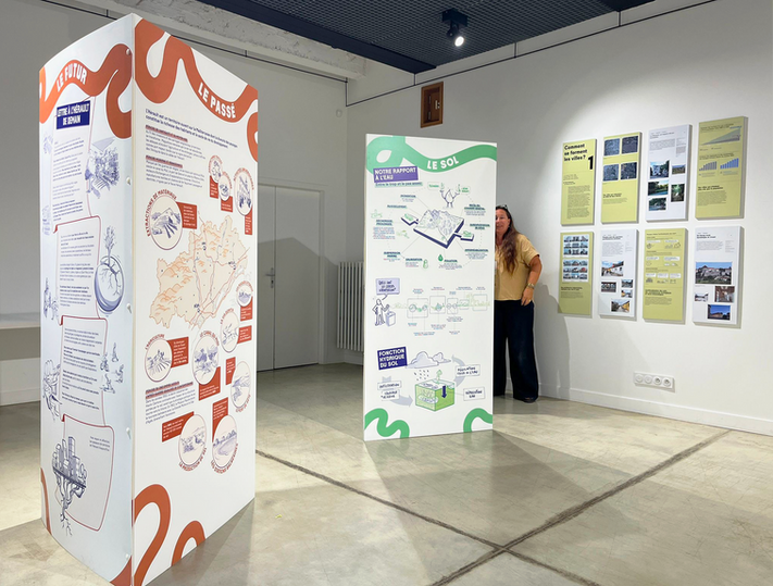 Photographie de deux panneaux d’exposition sur le thème du « Passé » et du « Sol ». Les structures verticales présentent des cartes et dessins explicatifs avec des titres colorés en rouge et vert. En arrière-plan, des panneaux muraux jaunes présentent d’autres contenus. Une personne se tient à droite de l’image.