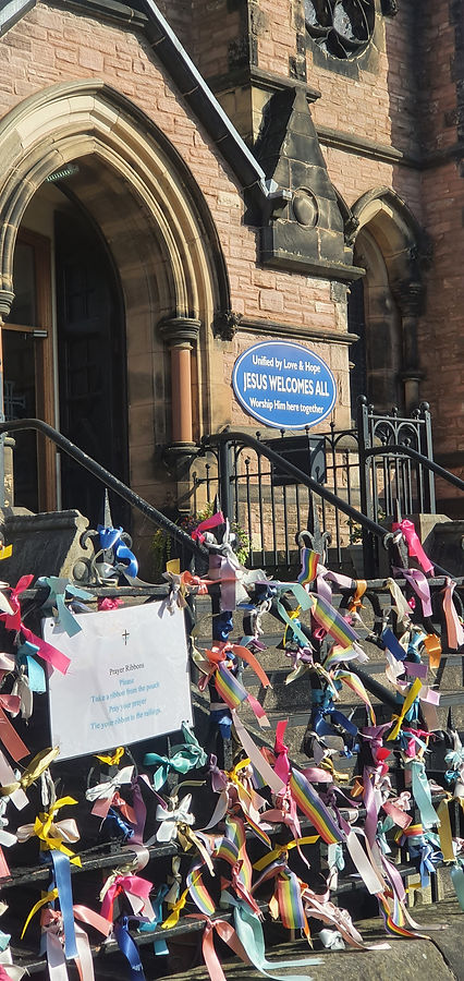 In the background is the entryway to a gothic church with a blue sign that reads "United by Love and Hope Jesus Welcomes All, Worship Him Here Together"
In the foreground are black iron railings with countless colourful ribbons tied on to them.