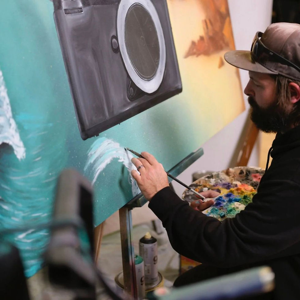 Artist painting a large camera on canvas, blending turquoise and white waves. Paint palette with multiple colors in the foreground.