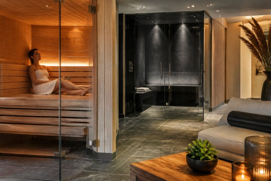 Hochwertige Wellness-Oase mit finnischer Sauna und Dampfbad in modernem Wohnbereich, warme Holzdetails, indirekte Beleuchtung, eine Person sitzt entspannt in der Sauna mit geschlossenen Augen