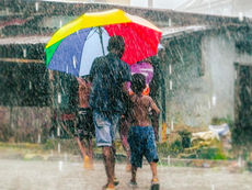 An adult and a child walk through heavy rain under a colorful umbrella on a flooded street in India during the monsoon season