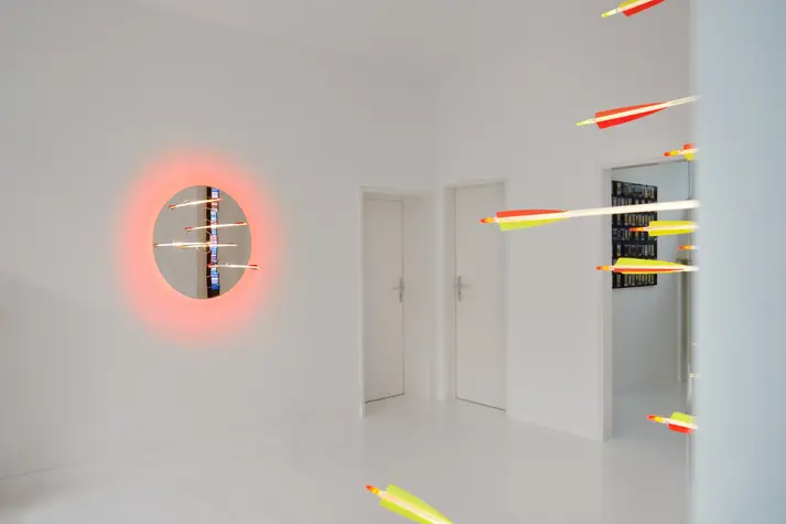 Weite weiße Galerieszene mit rundem Lichtkunstwerk von Hans Kotter an der Wand; mehrere leuchtende Pfeile scheinen von rechts durch den Raum zu fliegen.
