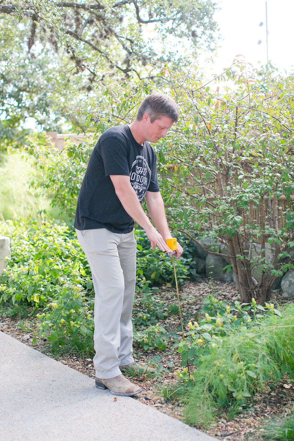 Man in a black T-shirt and beige pants measures soil moisture in a sunny garden, focused beside a yellow tool marker.
