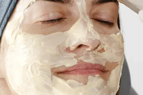 Person with a creamy facial mask relaxes with closed eyes, wearing a blue headband. Neutral background, conveying a calm mood.