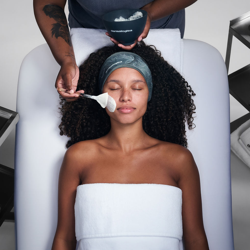 Person relaxing on a spa table, receiving a facial treatment with a brush. They're wrapped in a white towel. Text on bowl: Dermalogica.