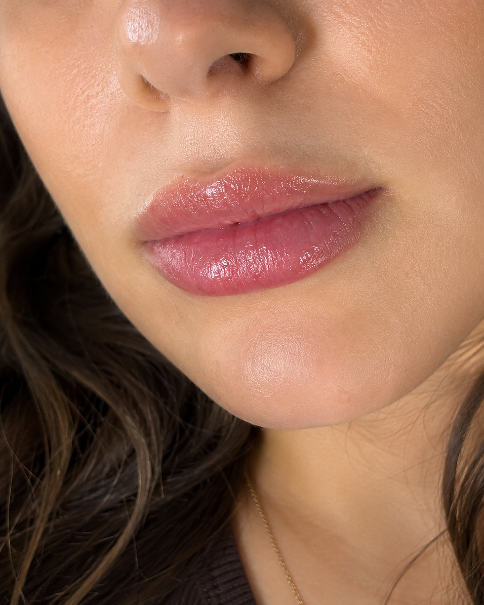Close-up of a woman's glossy pink lips and smooth skin. Dark hair frames her face, creating a soft and serene mood.