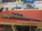 A rifle with a scope on a red workbench. Background shows tools, including screwdrivers and drill bits, organized on a workshop wall.