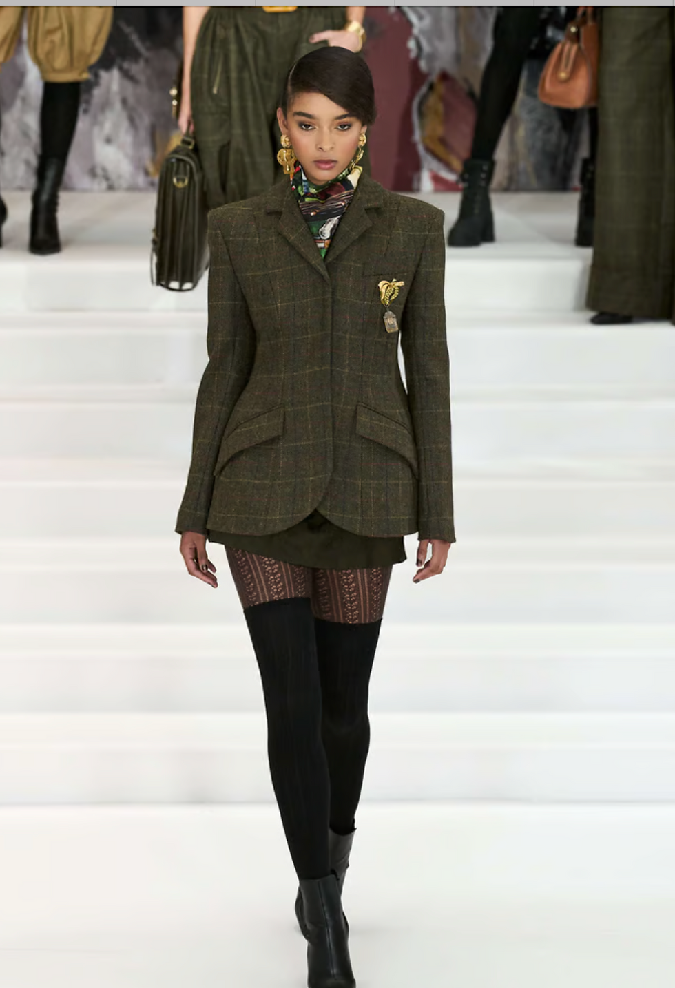 Model in a plaid blazer, patterned tights, and black boots walks on a white runway. Background shows more figures in similar attire. Fashion show.