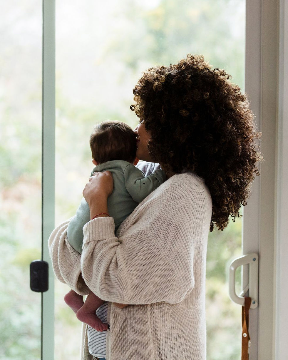 A woman standing by a window snuggling a baby.