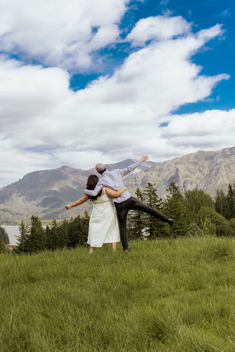 Couple standing in field with mountain view, arms outstretched, joyful pose.