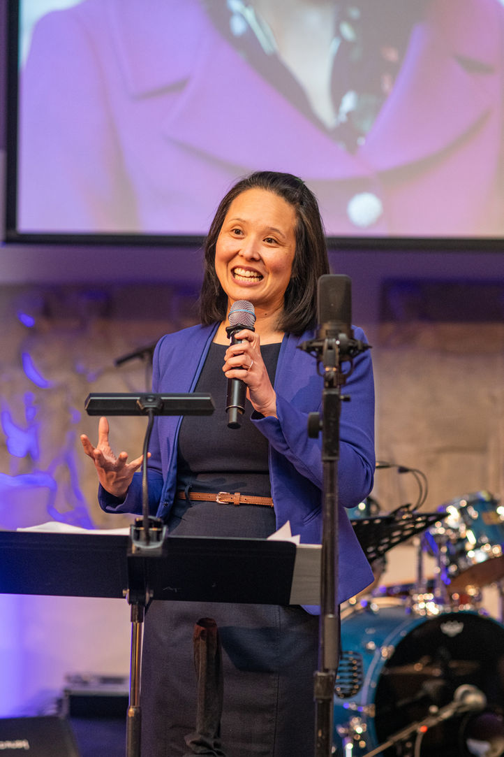 Smiling woman holding microphone speaking, wearing blue blazer, dark dress, and gesturing on stage.