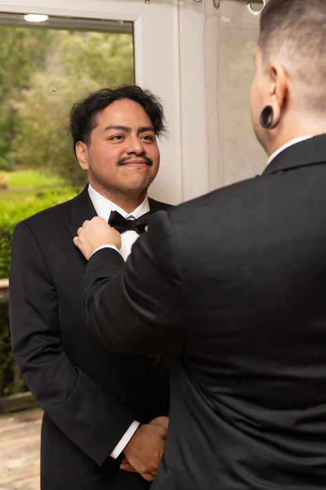 Smiling man in tuxedo gets bow tie adjusted by another.