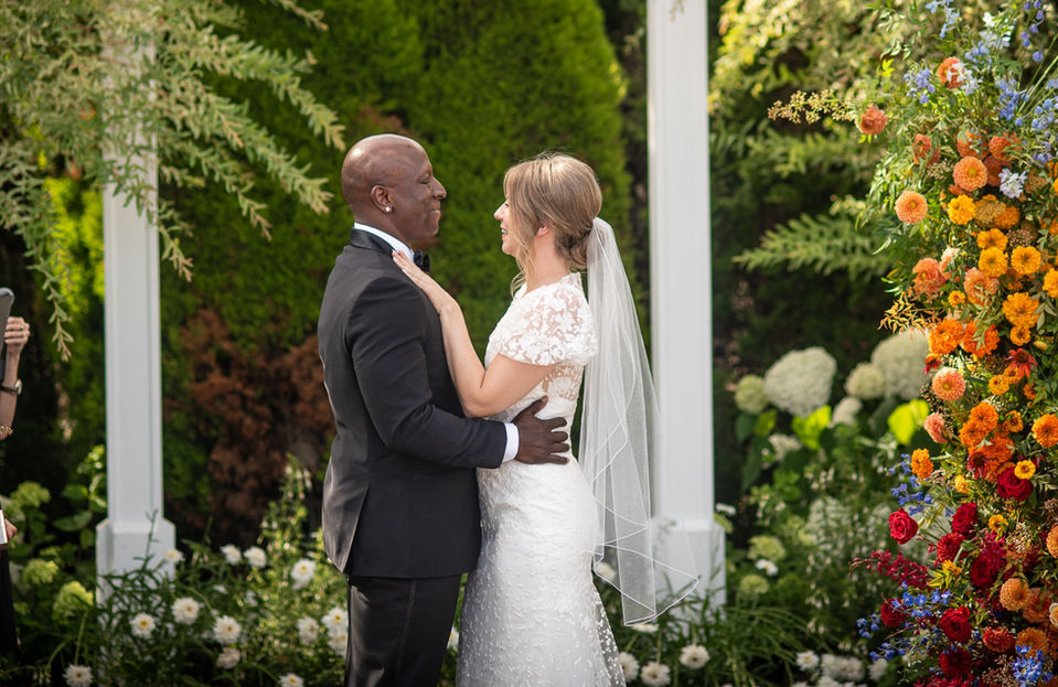 Couple at wedding ceremony, embracing with floral arch background.