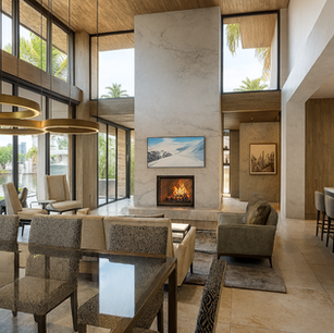 Contemporary double-height great room with floor-to-ceiling glass, marble fireplace wall, ring pendant lights, waterfront views, and open kitchen island seating.
