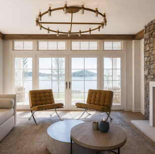 Lakefront living room with floor-to-ceiling French doors, exposed wood beams, stone fireplace, modern ring chandelier, and mid-century lounge chairs.