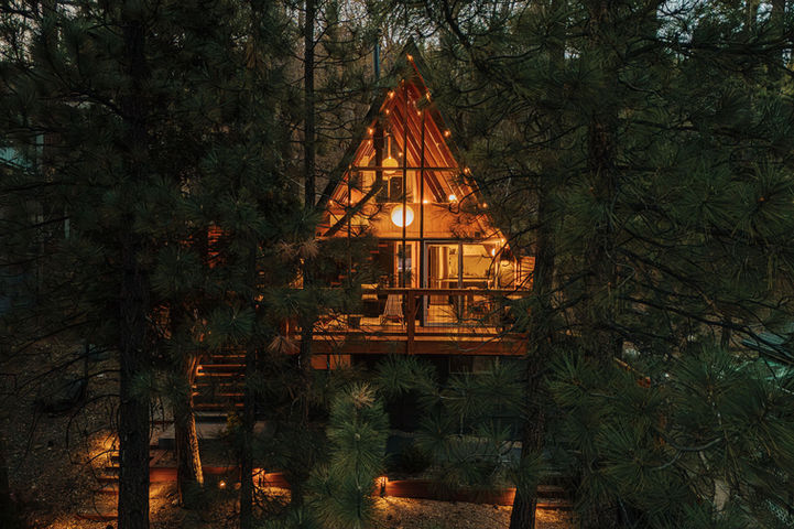 Twilight image of an A-frame in Big Bear Lake, CA. The warmly lit A-frame is framed by dark green pine trees. Photo by Gianna Christina Photo.