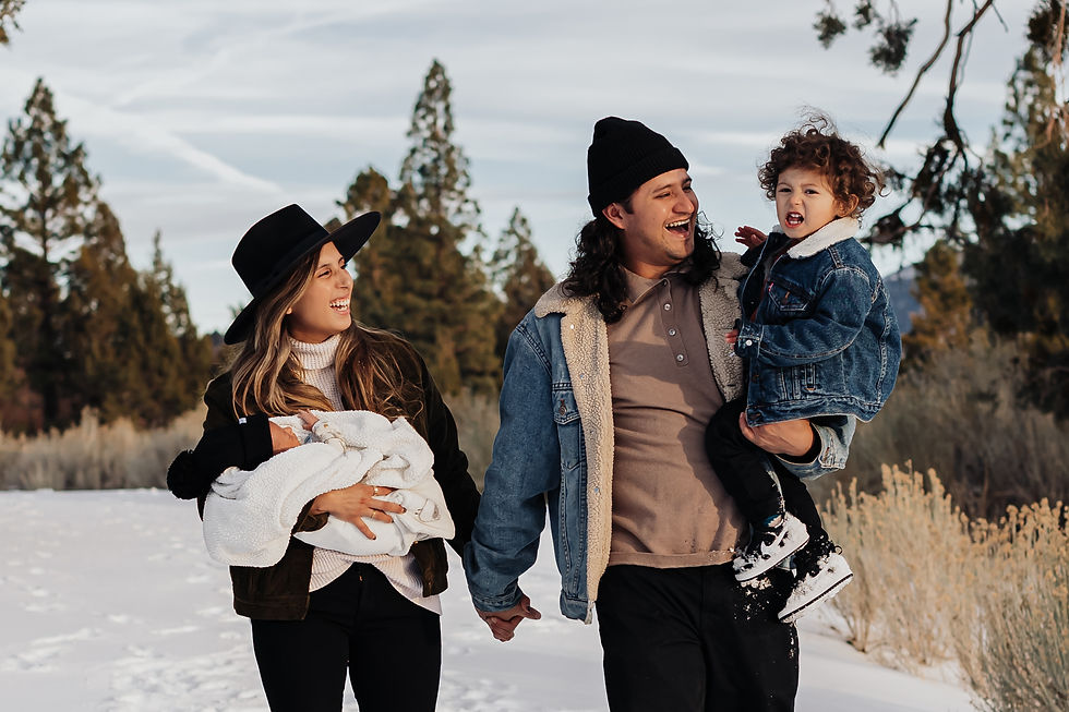 Hispanic family of four walking along a snow-covered path at Juniper Point in Big Bear Lake. There are pine trees in the distance. The dad and toddler are wearing blue jean jackets. They are all wearing black pants. The mom is wearing a black round hat, while the dad and newborn are wearing black beanies. Pops of golden sage bushes are to the right and behind them. Mom and dad are laughing at toddler who is screaming at the camera. It's an overcast day.