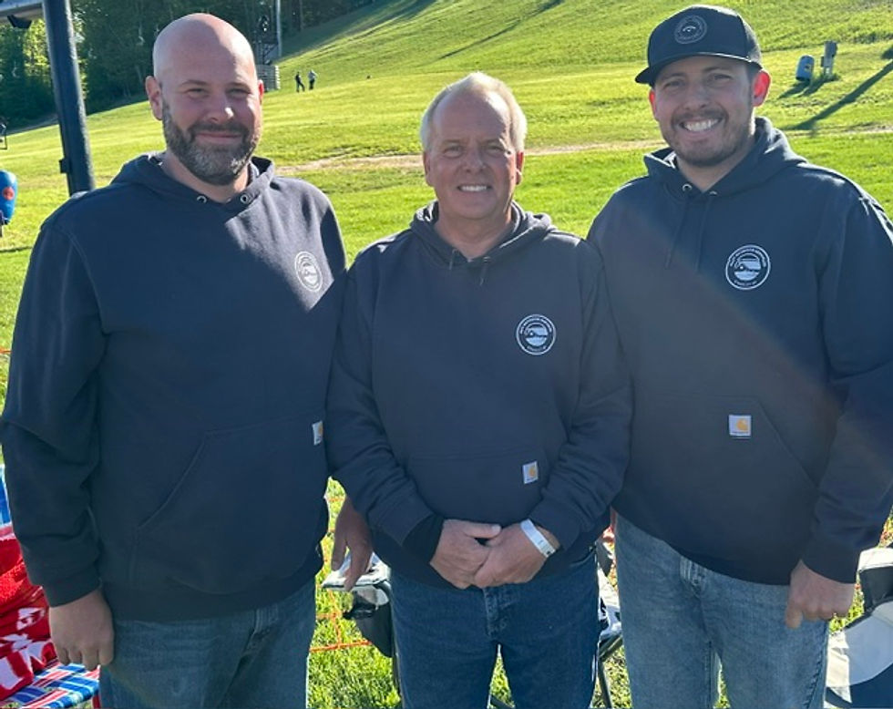 Outdoor photography of three men smiling side-by-side on a sunny, grassy field. They are dressed in matching casual hooded sweatshirts, creating a friendly and collegiate atmosphere.