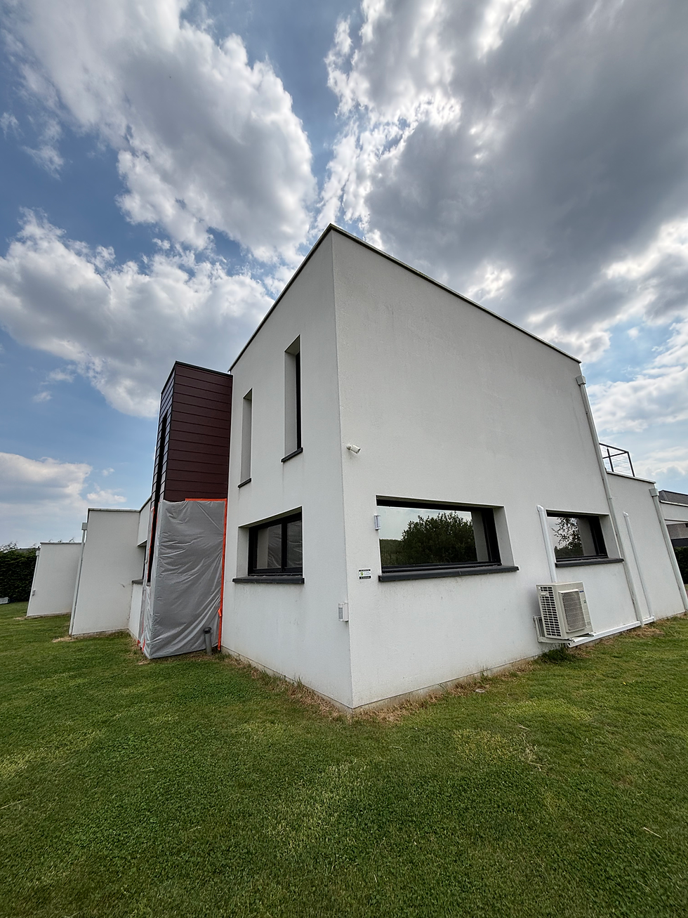 Maison moderne blanche sur pelouse verte, ciel nuageux. Mur avec fenêtres teintées, unité de climatisation extérieure visible. Atmosphère paisible.