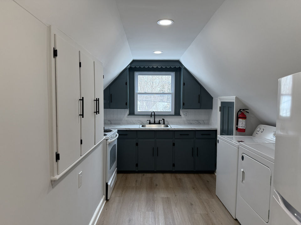 Updated kitchen in Ridgewood NJ rental apartment featuring hardwood floors, sloped ceiling, dark lower cabinets, white appliances, in-unit washer and dryer, and natural light from a central window.
