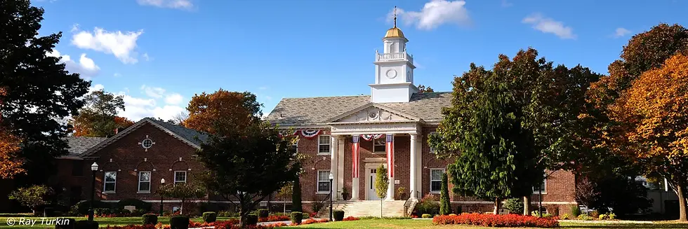 Historic red brick municipal building in Teaneck, NJ with white columns, American flags, fall foliage, and a clear blue sky. Great for showcasing homes for sale in Teaneck, NJ, real estate near top Bergen County schools, and Northern NJ properties in established neighborhoods.