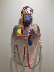 A sculpture made of yarn on a plastic frame. The frame is shaped like a human torso and the yarn is many colors, with pink being dominant. A life-size crochet heart and crochet string of guts hang from the frame.