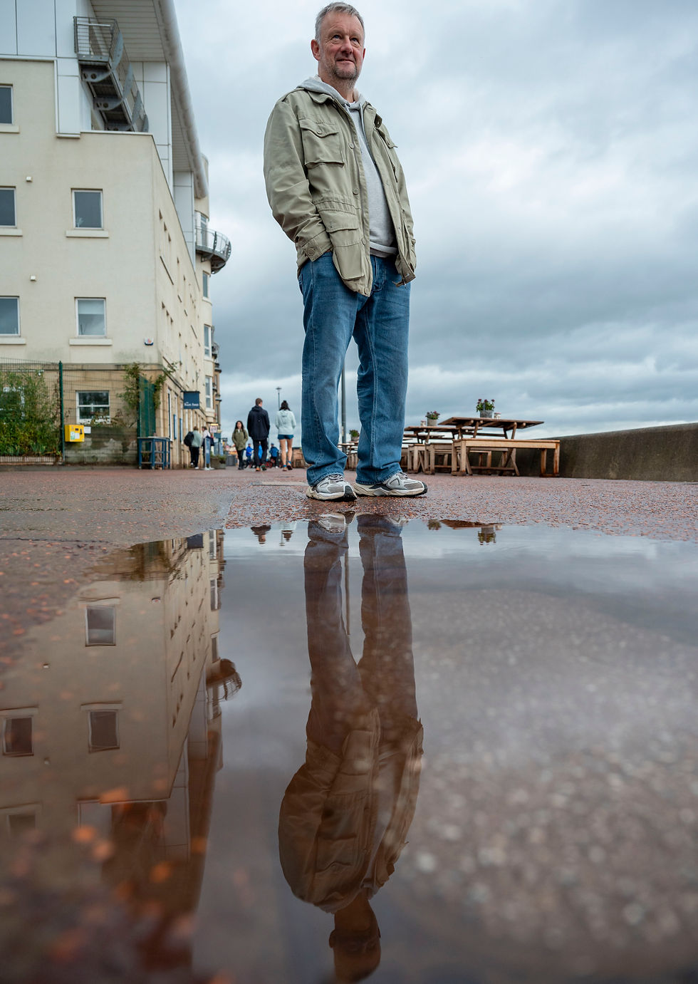 Man stands in puddle, reflection visible, overcast sky. Public figure in Portobello.
