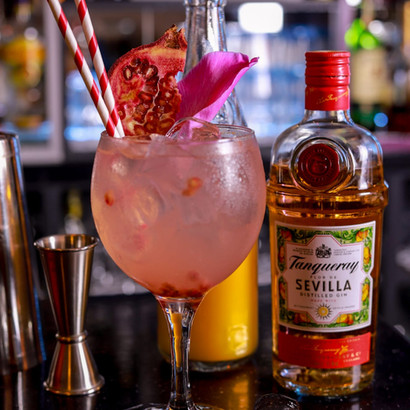 Pink cocktail with garnish, two straws, on bar with gin bottle.