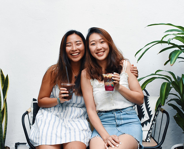 Two smiling women, co-founders of An Açaí Affair, sitting side-by-side on a bench against a white wall, holding drinks and enjoying each other’s company, with green plants in the background