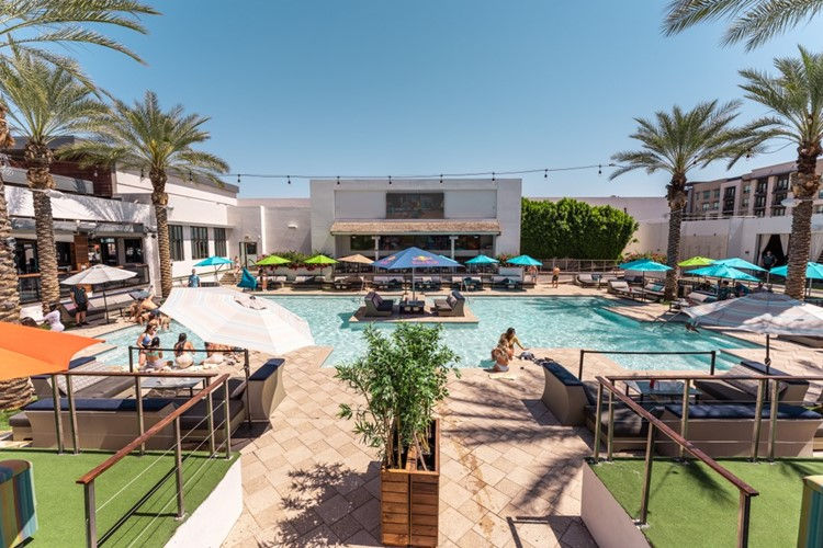 Pool scene with people lounging, surrounded by palm trees and colorful umbrellas. Clear sky and modern building in background.