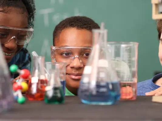 Three students wearing safety goggles examine colorful liquids in beakers, smiling in a science classroom with a chalkboard background.
