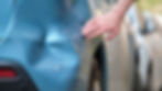 Hand touching a dented blue car door after a crash, with scratched paint and a blurred parked vehicle in the background.